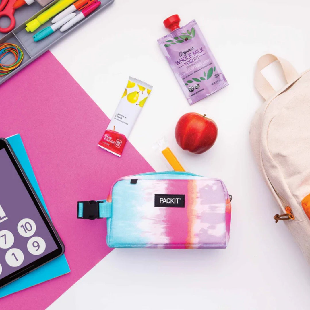 A PACKIT brand zippered pencil case with a pastel tie-dye pattern in blue, pink, and purple, styled in a flat-lay school supply arrangement on a white and pink background.