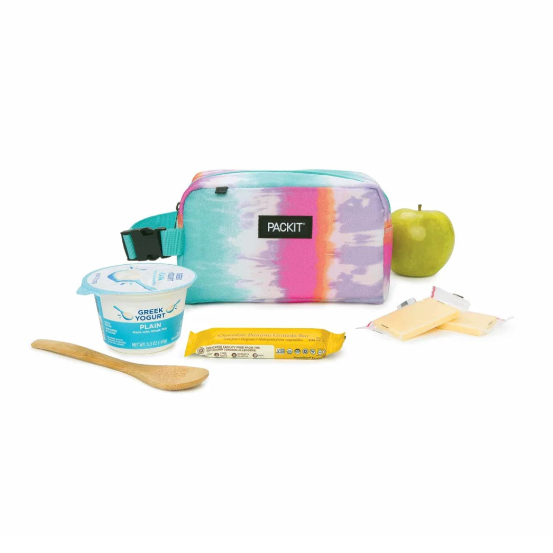 A Packit lunch bag in a pastel tie-dye pattern, shown with a Greek yogurt cup, a wooden spoon, a sandwich, cheese slices, and an apple on a white background.