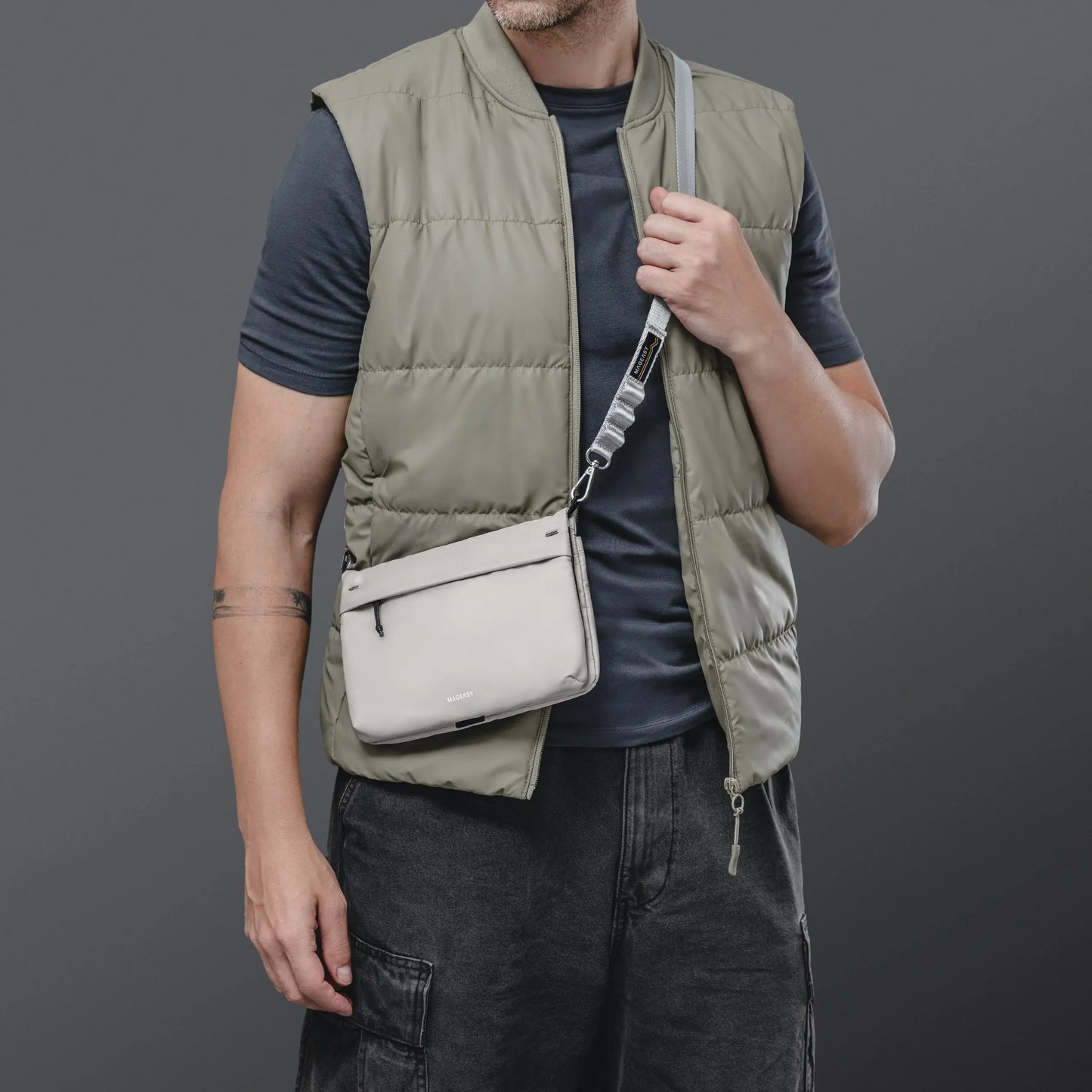 A man wearing a light olive green puffer vest over a dark gray t-shirt and dark gray cargo pants, carrying a light beige crossbody bag with a gray strap.