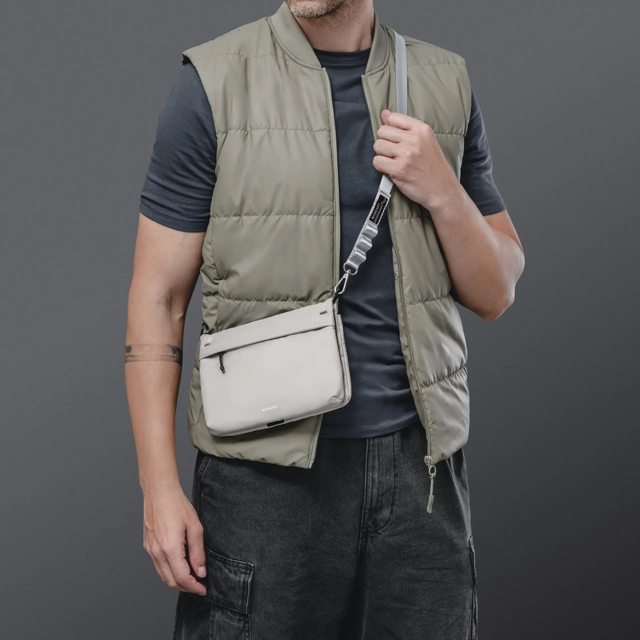 A man wearing a light olive green puffer vest over a dark gray t-shirt and dark gray cargo pants, carrying a light beige crossbody bag with a gray strap.
