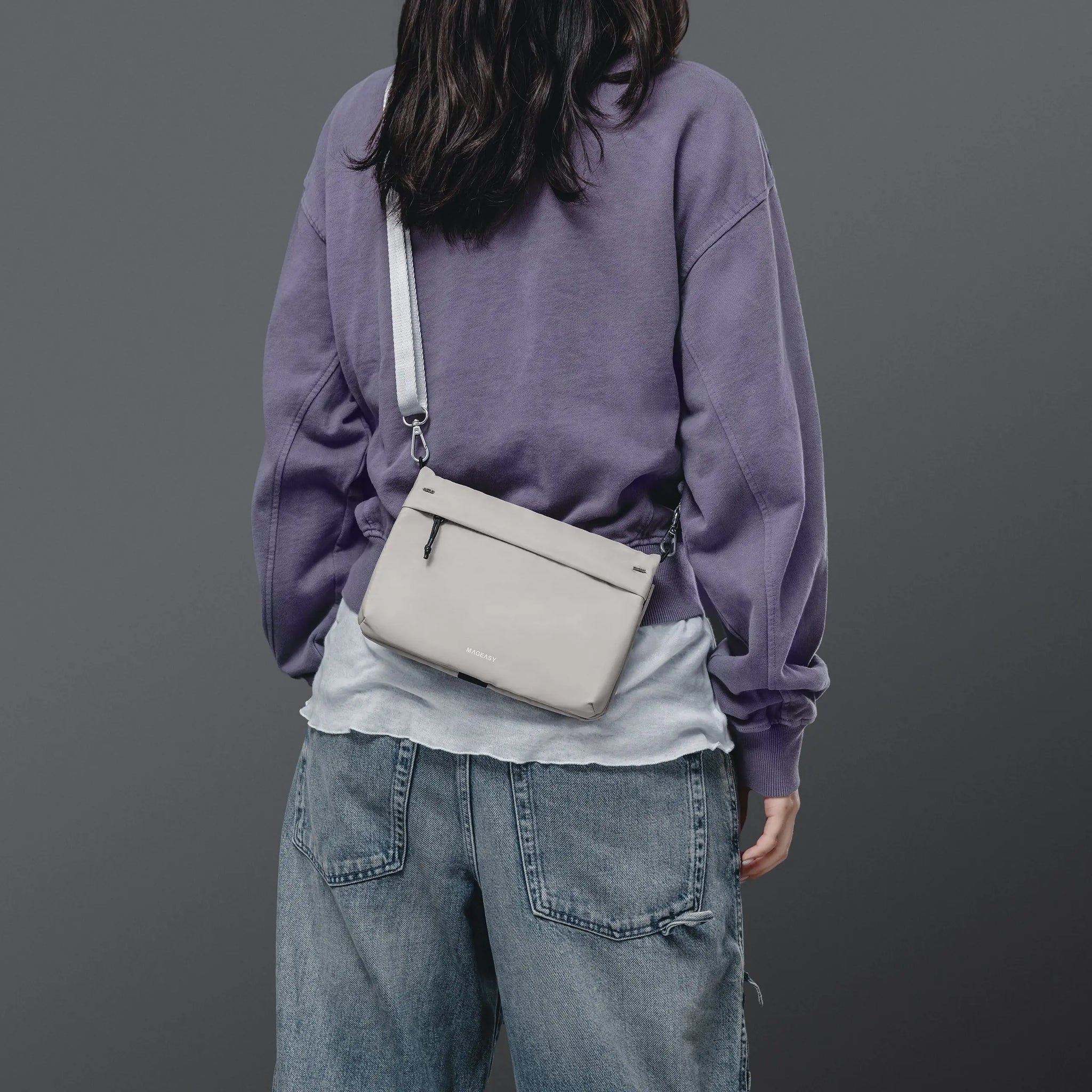 A person shown from the back wearing a lavender sweatshirt and light blue jeans, carrying a light gray crossbody bag with a white strap against a dark gray studio background.