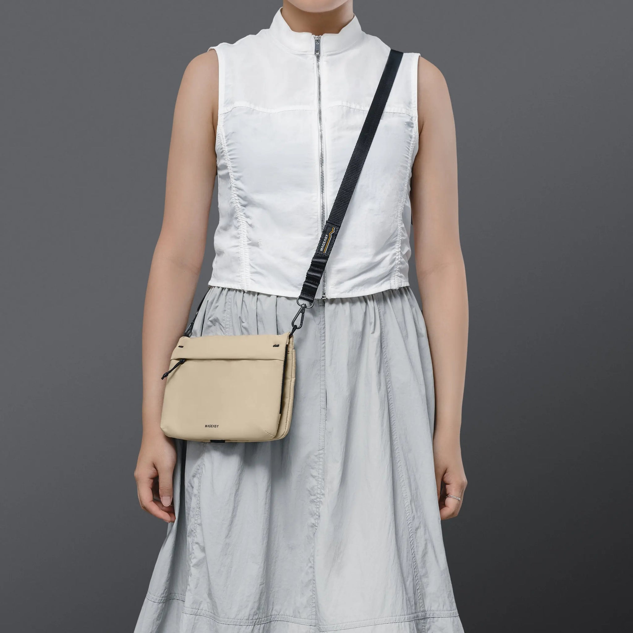 A model wearing a white sleeveless top and a light gray skirt carries a beige crossbody bag with a black strap against a dark gray studio background.