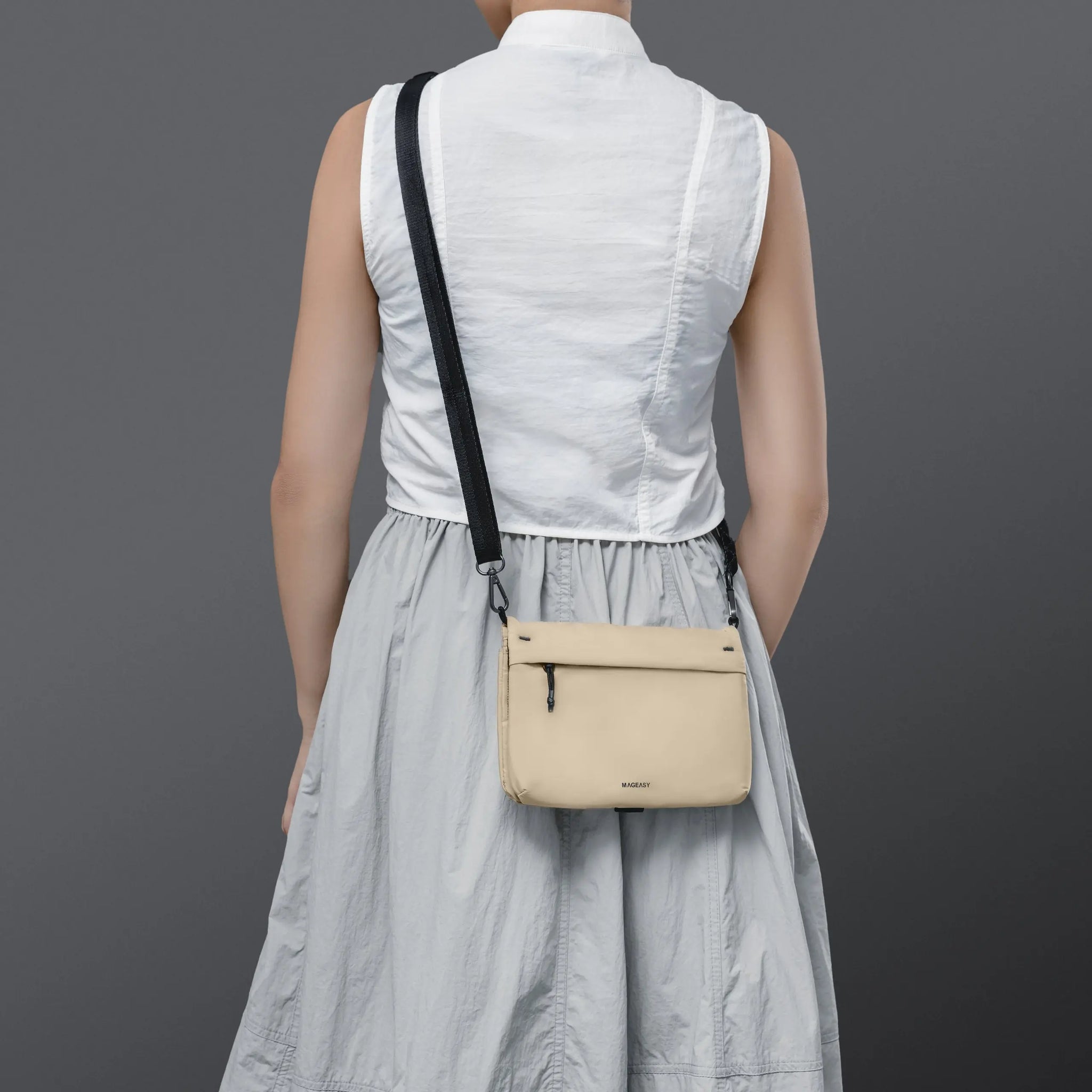 A person shown from the back wearing a sleeveless white top and light gray skirt, carrying a small beige crossbody bag with a black strap against a dark gray studio background.