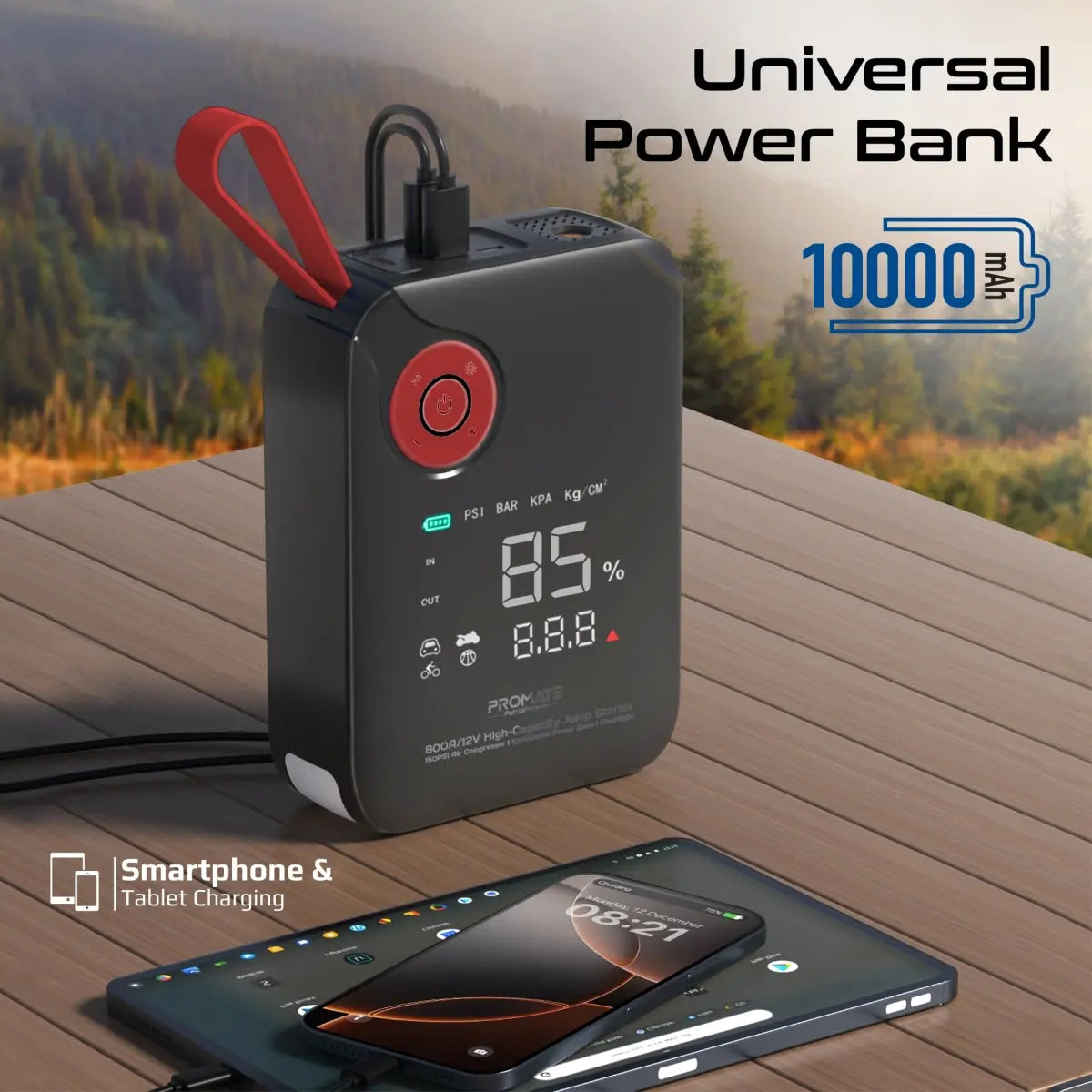 A black portable power bank with a red carrying loop and digital display, shown on a wooden table outdoors with a blurred forest background. A smartphone and tablet are charging from its USB-C port.