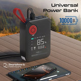 A black portable power bank with a red carrying loop and digital display, shown on a wooden table outdoors with a blurred forest background. A smartphone and tablet are charging from its USB-C port.